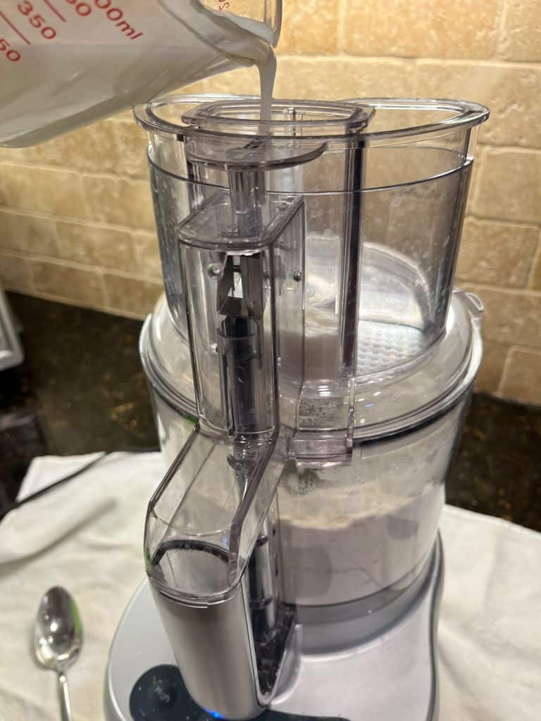 Milk being poured down food processor chute while running to mix with dry ingredients.