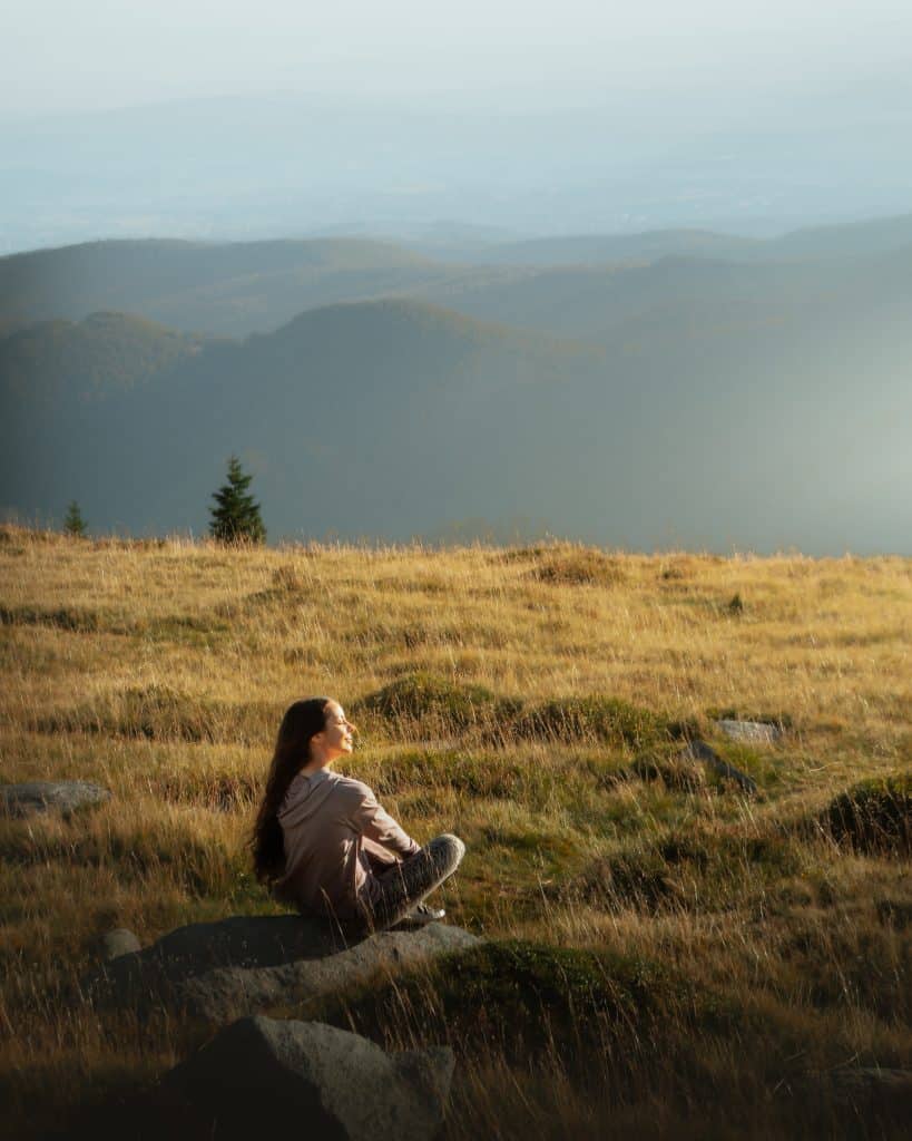 Lady sitting in field with eyes closed, relaxing with mountains in background